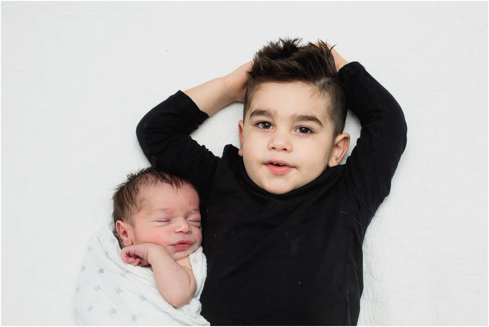 Toddler relaxing with his hands above his head next to his newborn baby brother.