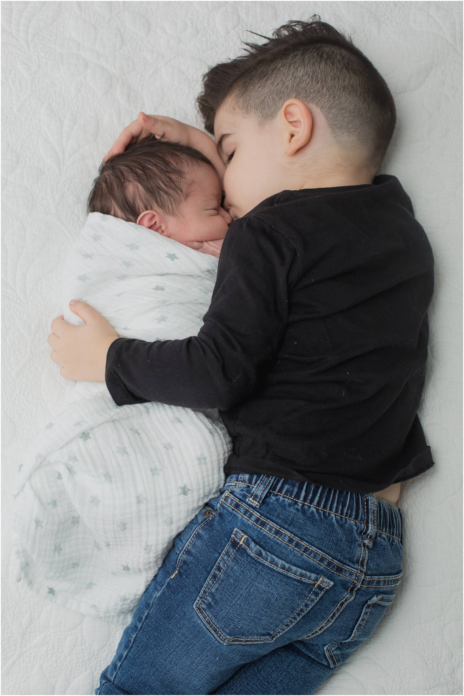 Toddler lies on bed and snuggles his newborn baby boy in neutral swaddle during lifestyle newborn session.