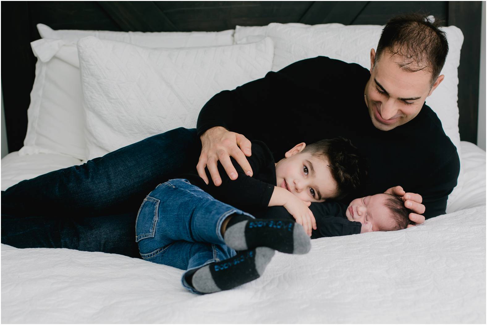 Dad snuggles newborn baby boy and toddler on their bed all wearing black tops.