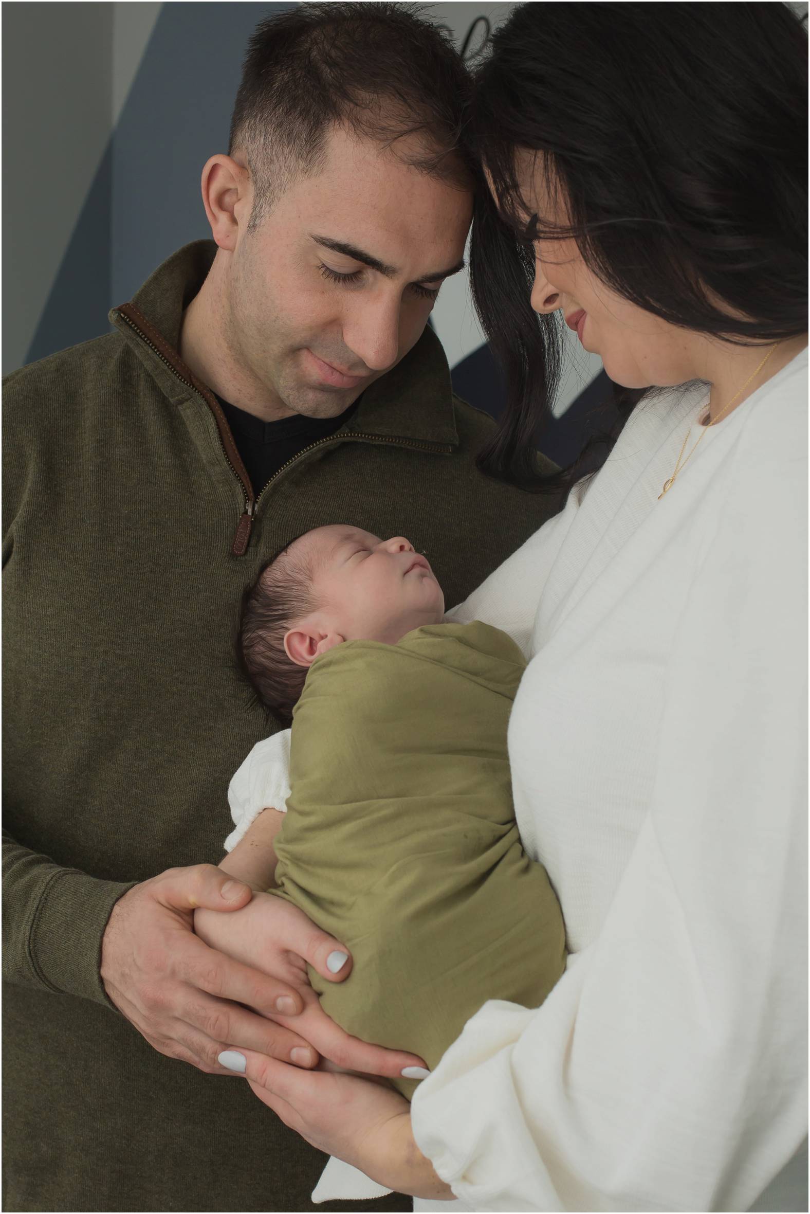 Mom and dad snuggle newborn swaddled in olive green blanket