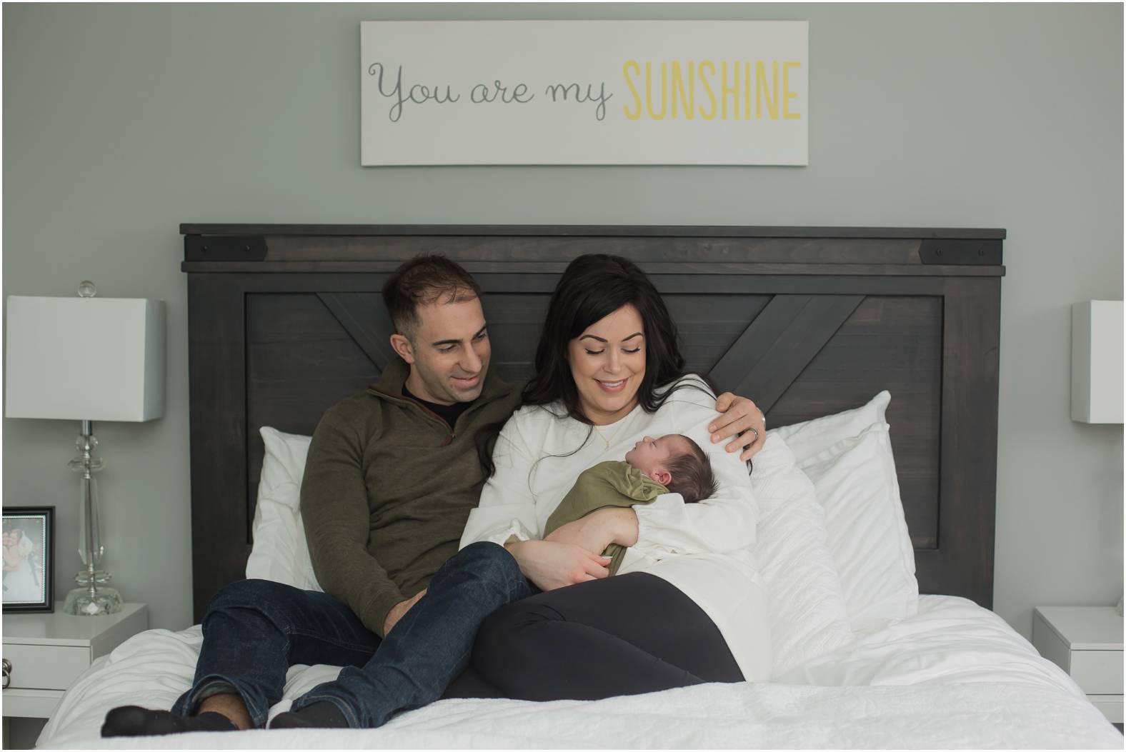 Parents snuggle newborn baby boy swaddled in olive green blanket on the bed.
