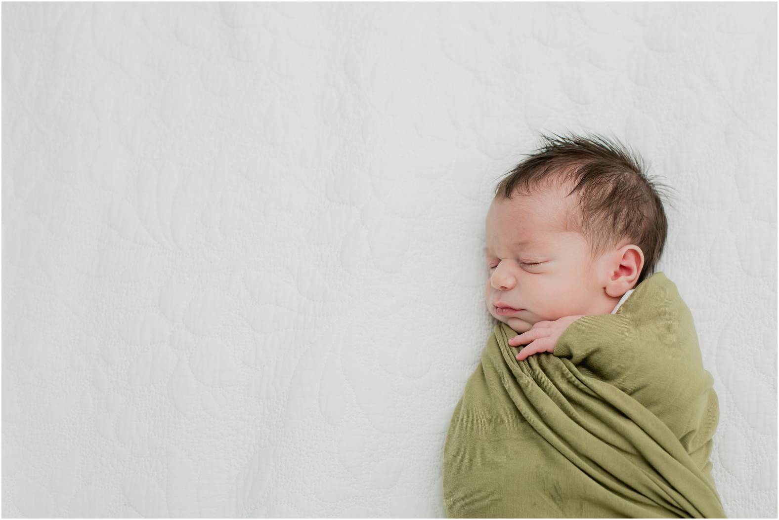 Newborn baby boy swaddled in olive green blanket poses for newborn photoshoot done in the home.