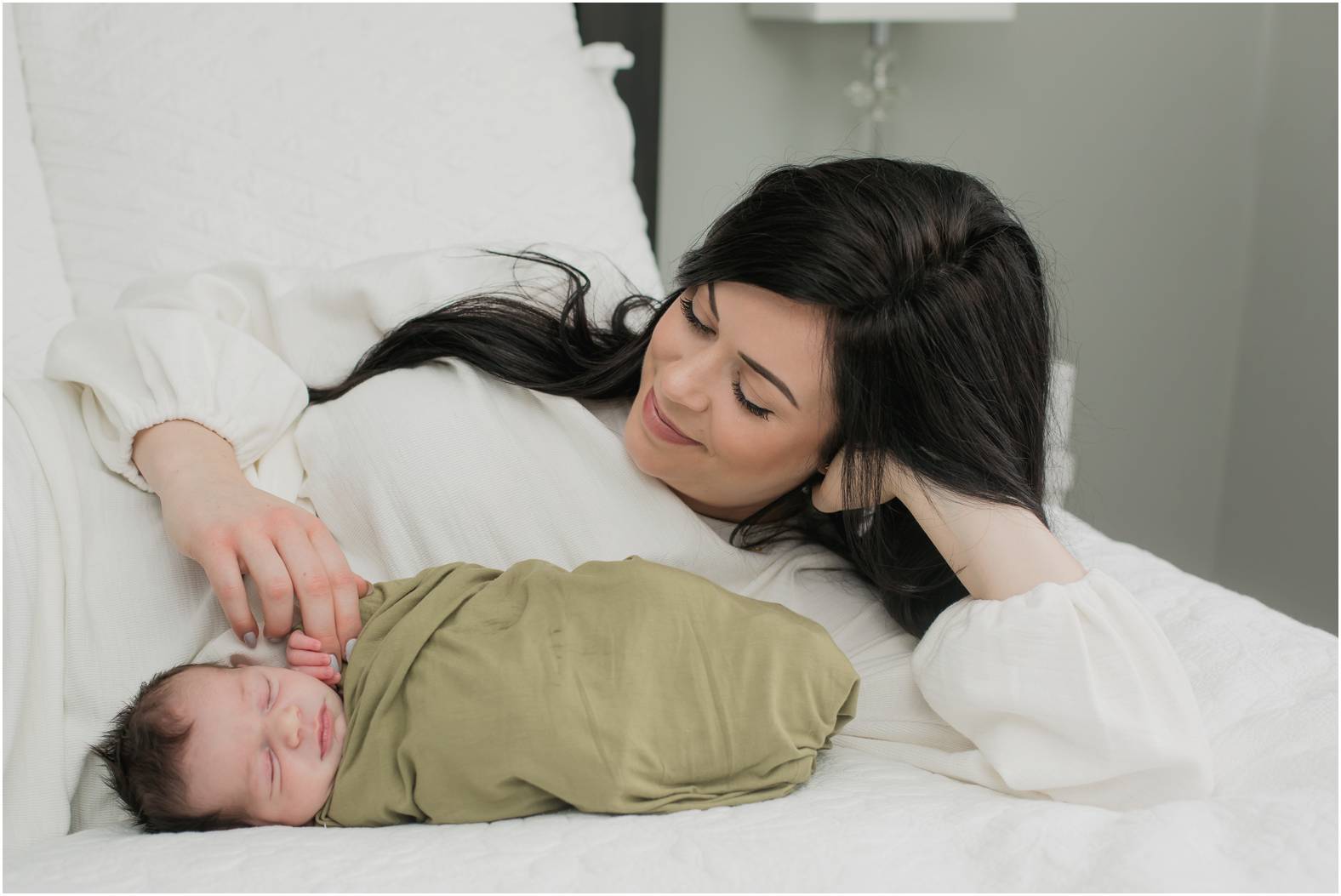 Beautiful new mom snuggles newborn baby boy swaddled in an olive green blanket on the bed.