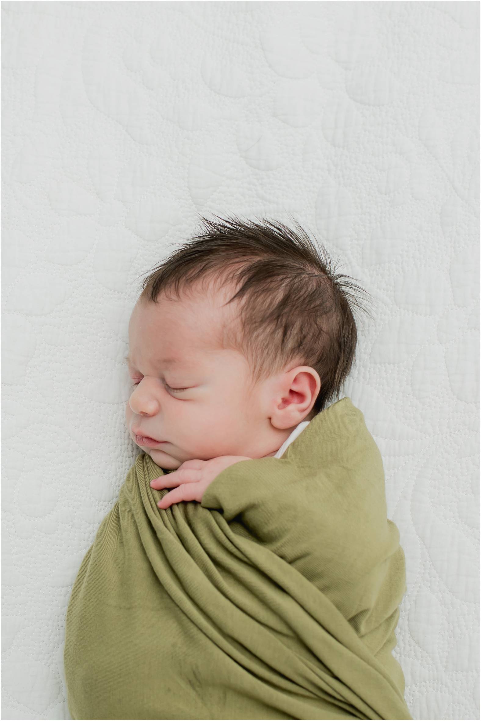 Newborn baby boy swaddled in olive green blanket poses for newborn photoshoot done in the home.